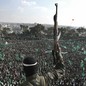 A militant, photographed from behind, holds a gun in the air as he stands before a crowd waving green flags